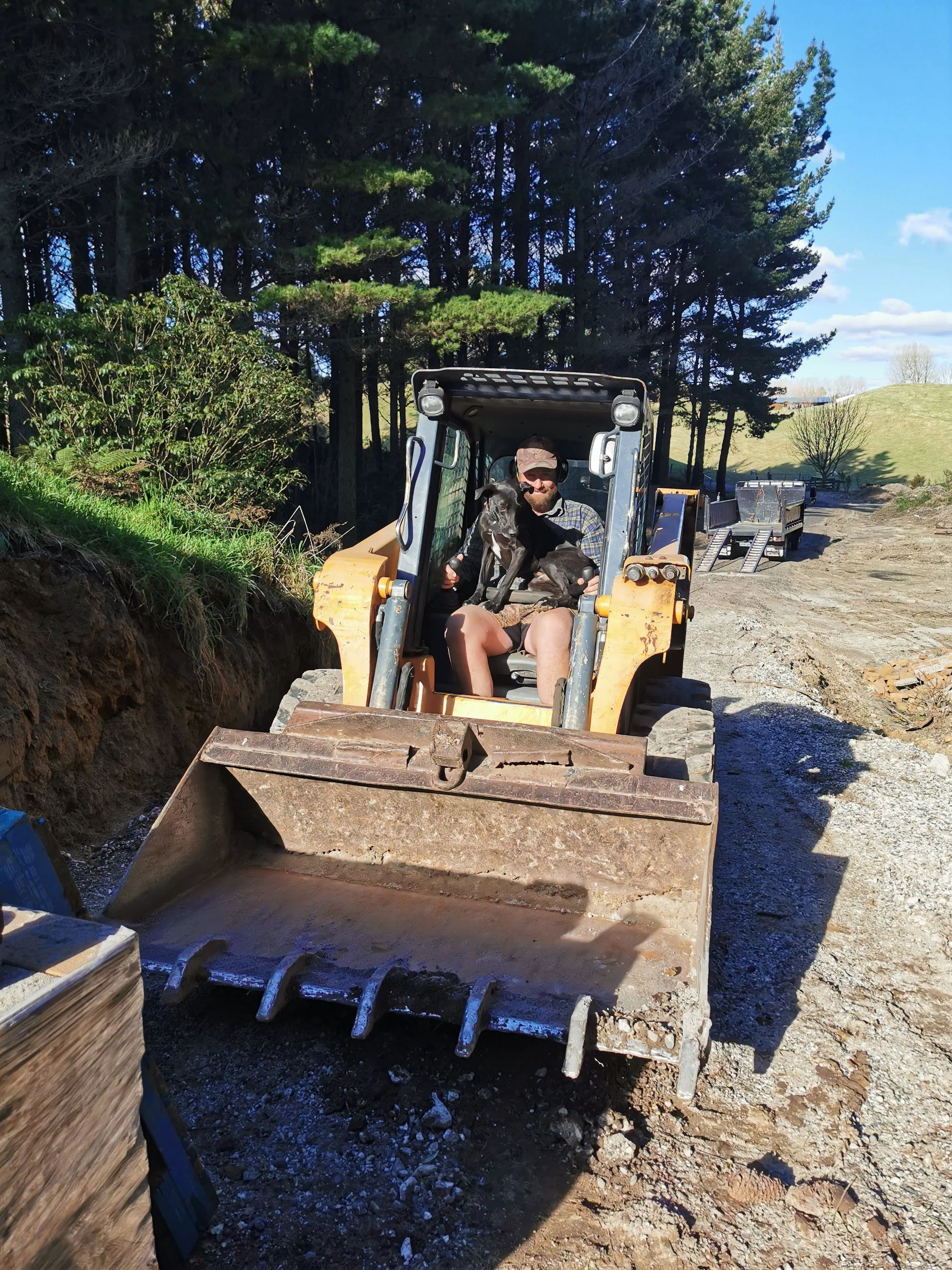A man operating a bulldozer with a black dog on his lap, on a construction site with trees and a partly cloudy sky in the background.