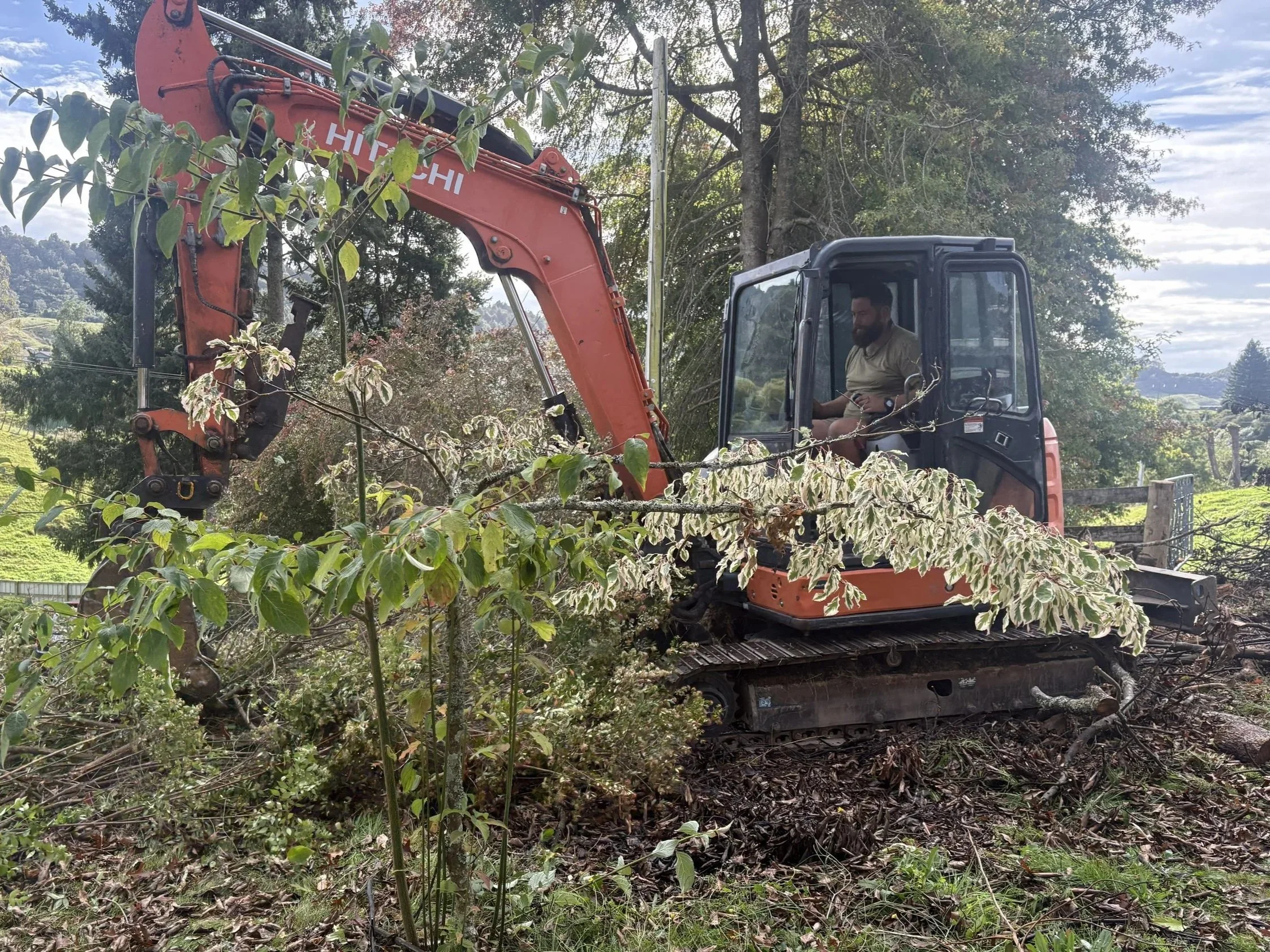 A person operating a small orange and black excavator on a grassy, wooded landscape with trees and rolling hills in the background.