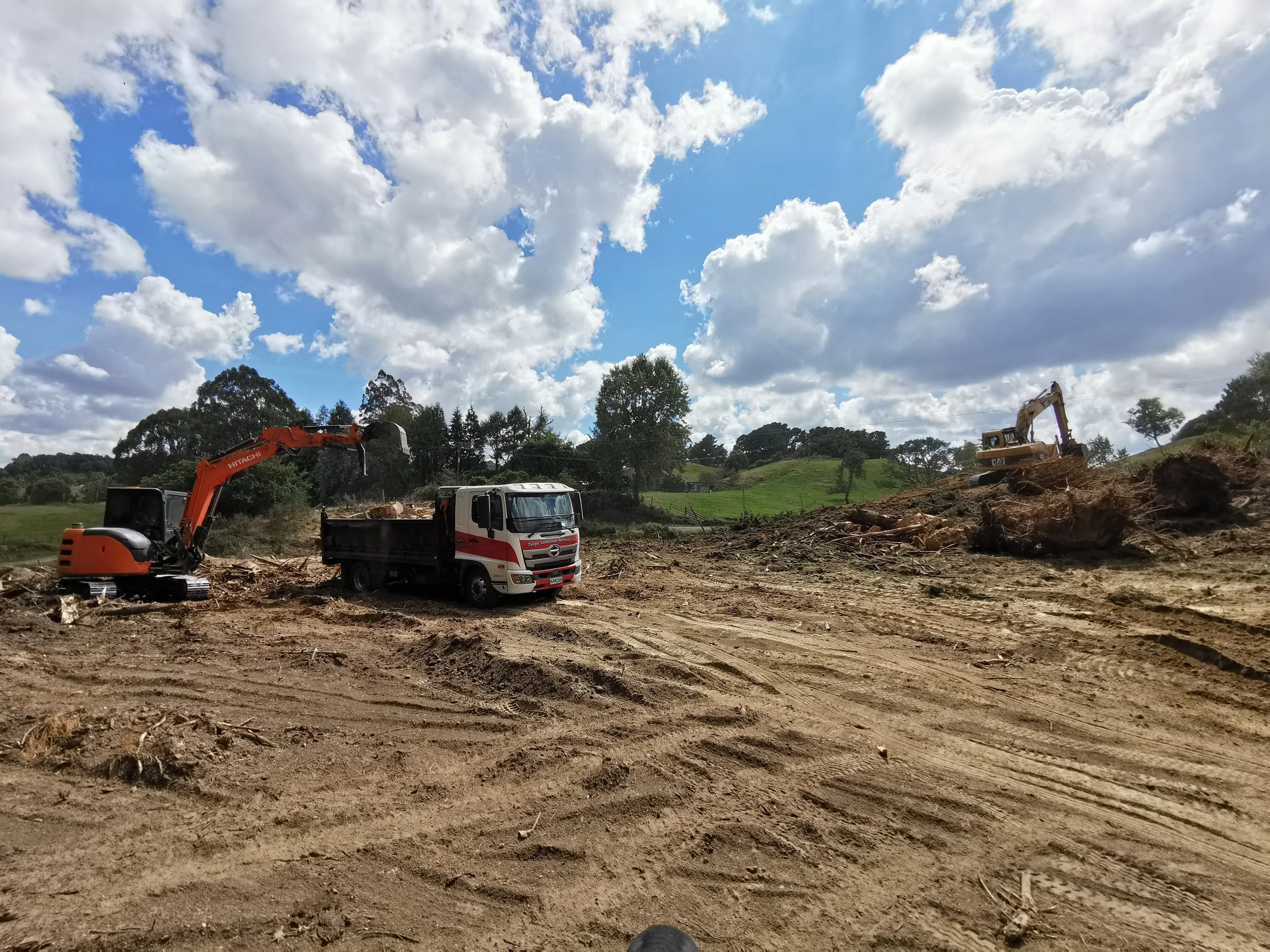 Construction site with excavators and a truck working on the land, surrounded by green hills and a partly cloudy sky.