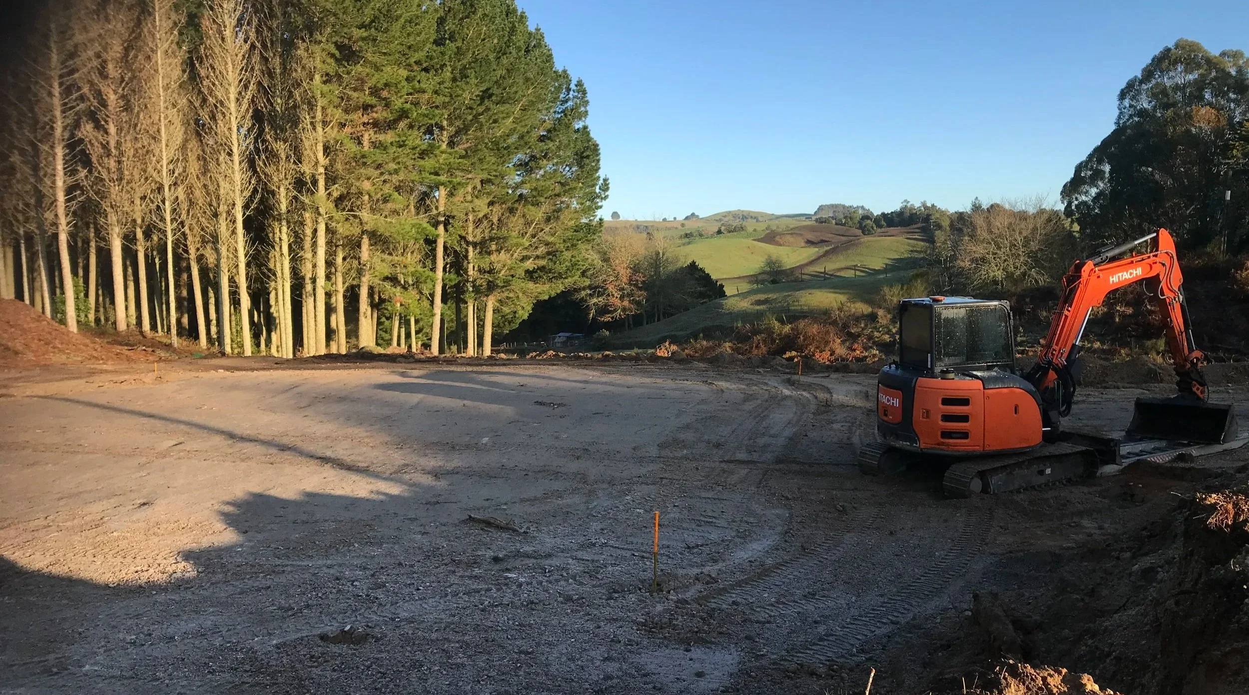 Construction site with a small orange excavator on dirt ground, surrounded by trees and rolling hills in the background.