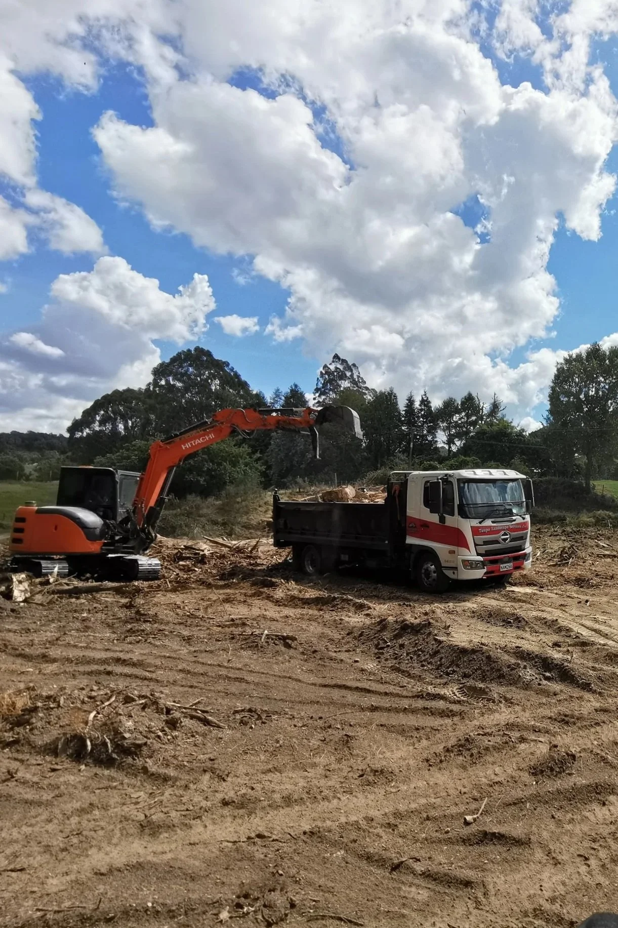 An orange excavator loading dirt into a white and red dump truck on a construction site under a blue sky with scattered clouds.