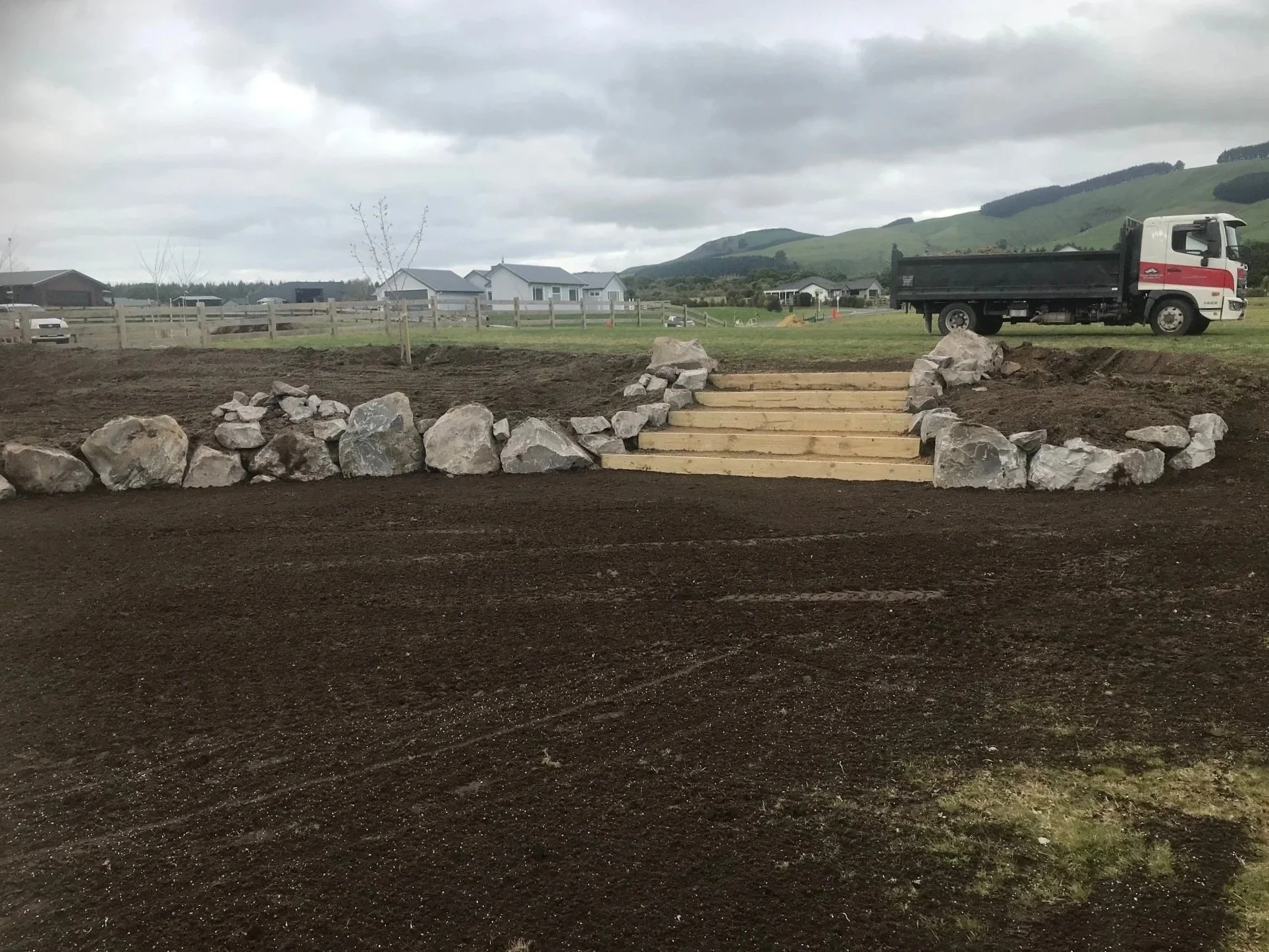 Construction site with a wooden stairway surrounded by large rocks, dirt ground, and a truck in the background, with residential homes and green rolling hills under cloudy skies.