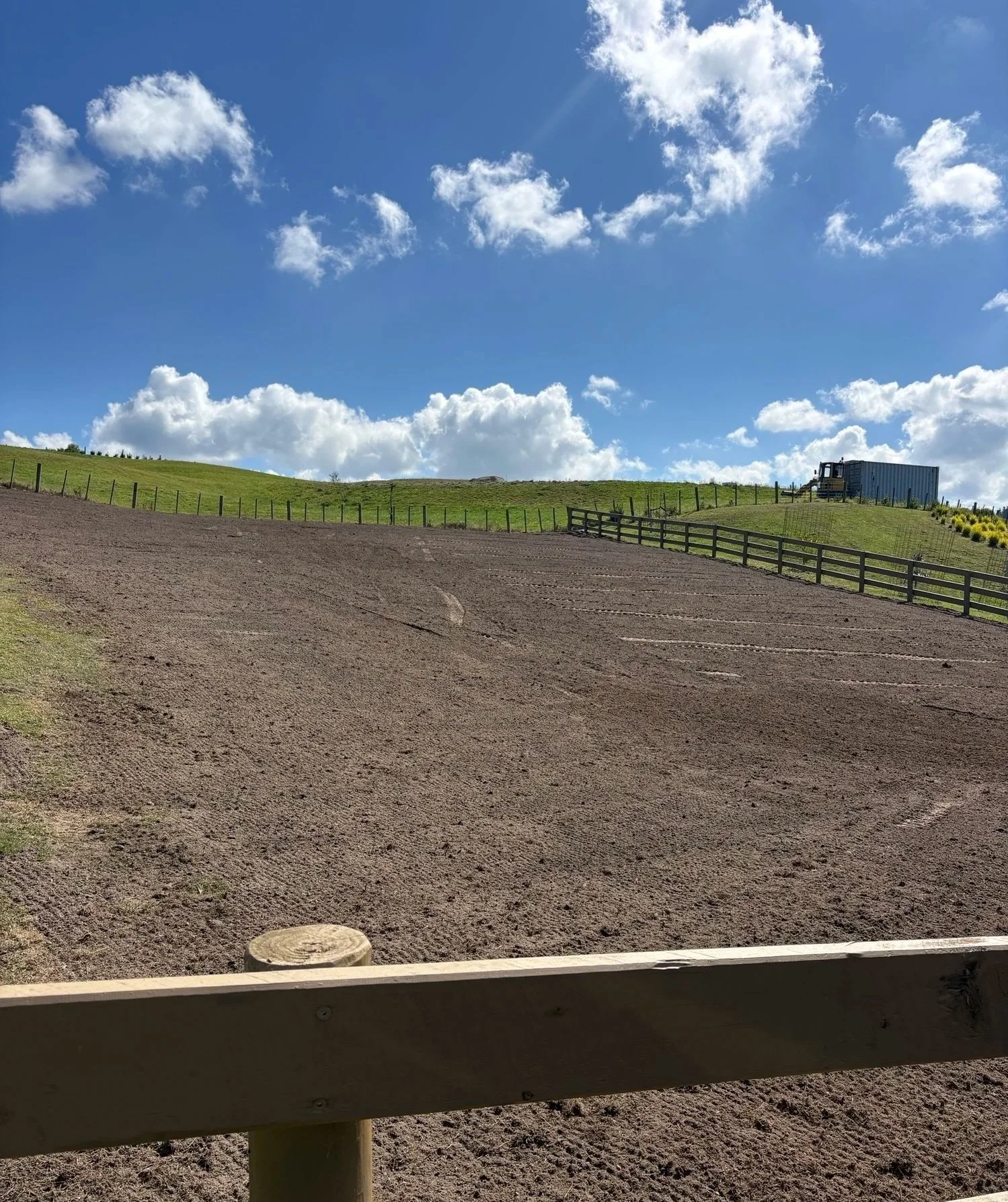 A dirt slope surrounded by a wooden fence, green grass on top of the hill, and a blue truck or container near the top right on a sunny day with a blue sky and scattered clouds.
