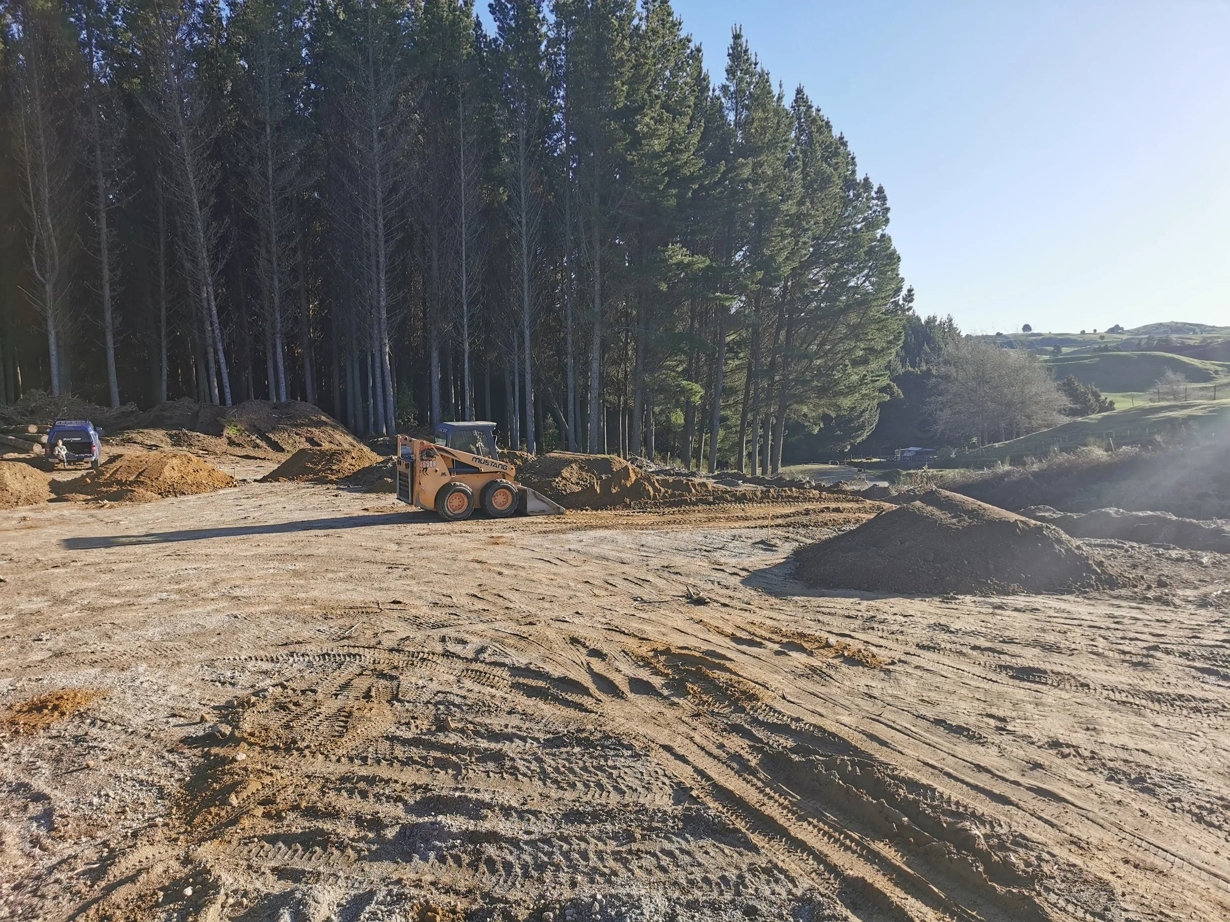A construction site with a bulldozer, mounds of dirt, and a truck, set against a backdrop of trees and rolling hills under a clear sky.