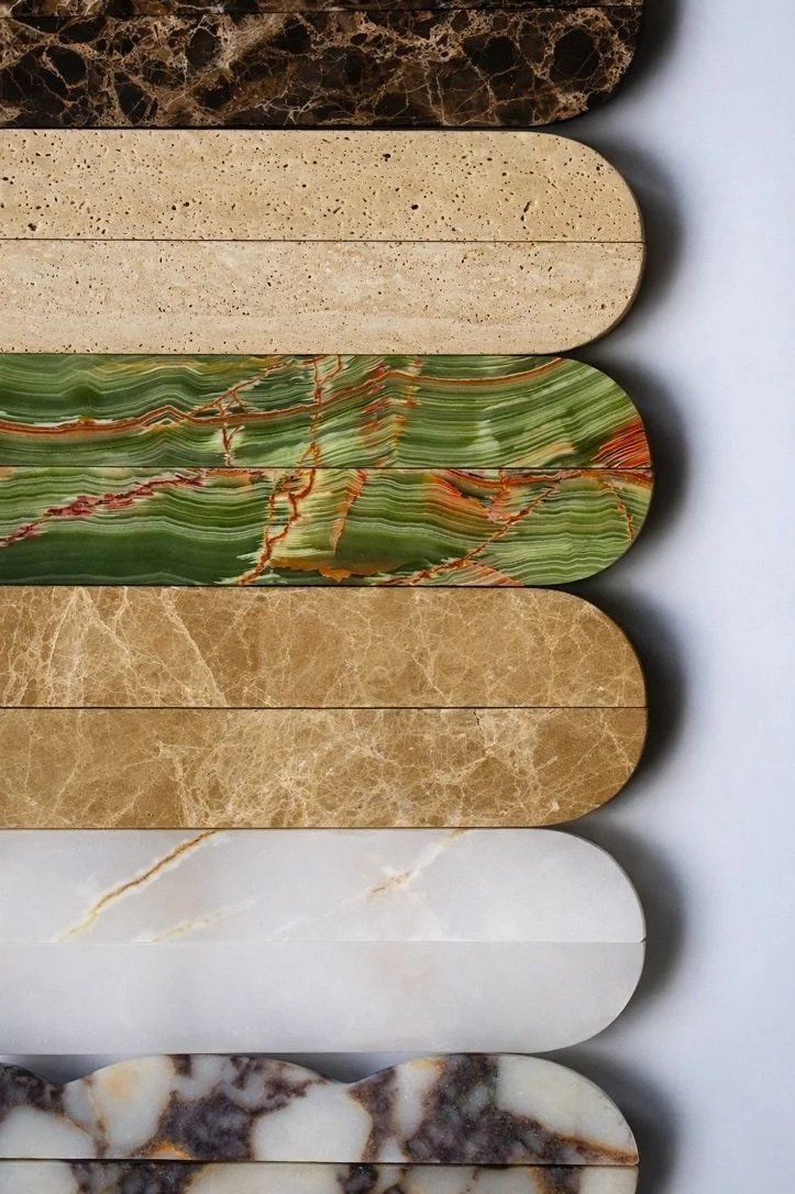 A selection of six different marble and stone samples arranged horizontally. The top sample is dark brown with a marble pattern, the second is beige with small dark speckles, the third is green with red and orange veins, the fourth is light brown with a marble-like texture, the fifth is white with subtle veins, and the bottom sample is white and dark brown marble with a marbled pattern.