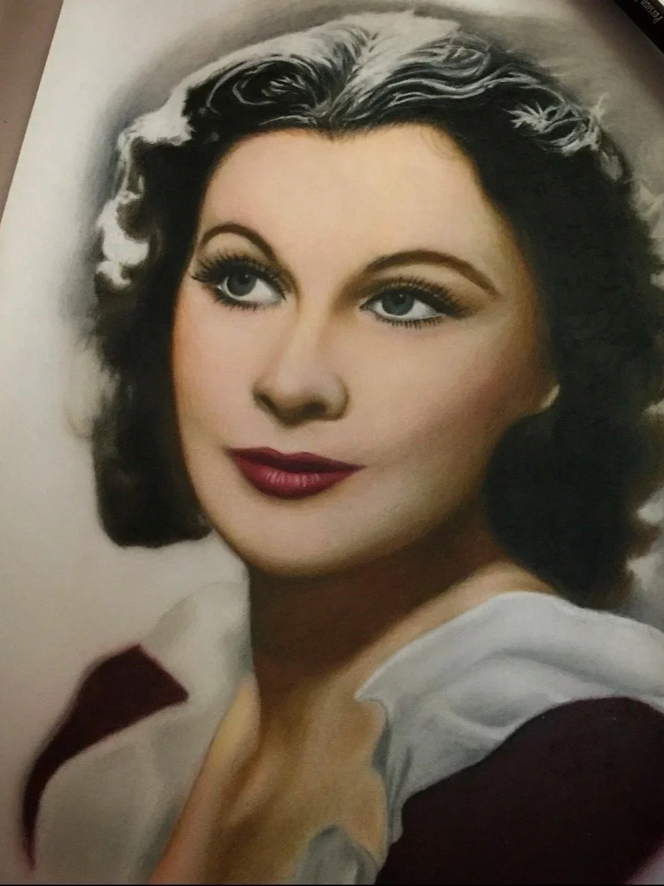 Close-up photograph of a woman's face with dark, wavy hair, blue eyes, red lipstick, and wearing a white and maroon outfit.