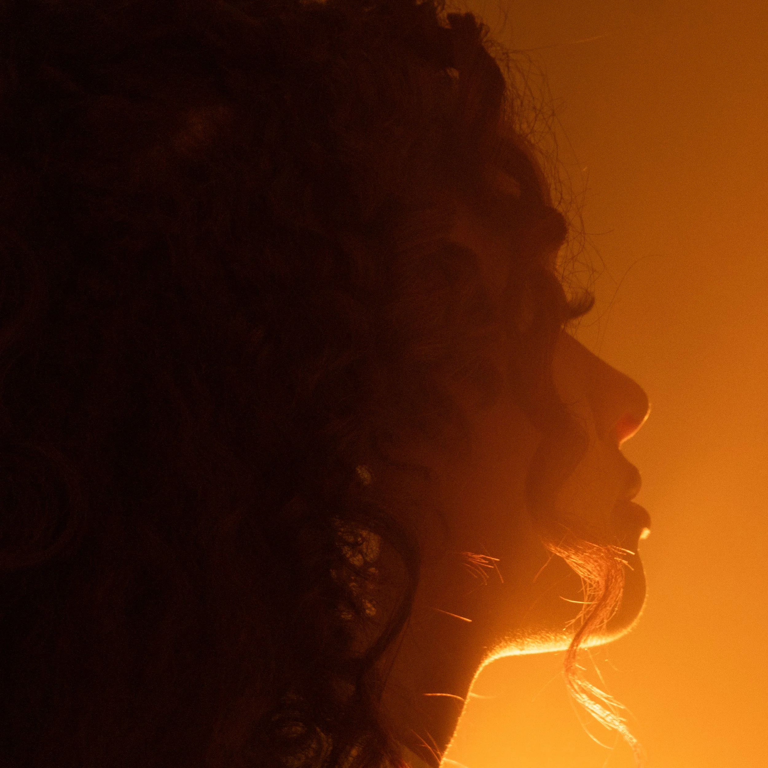 Silhouette d'une personne avec un fond orange de feu ou de lumière intense, visage partiellement visible, cheveux longs et ondulés.