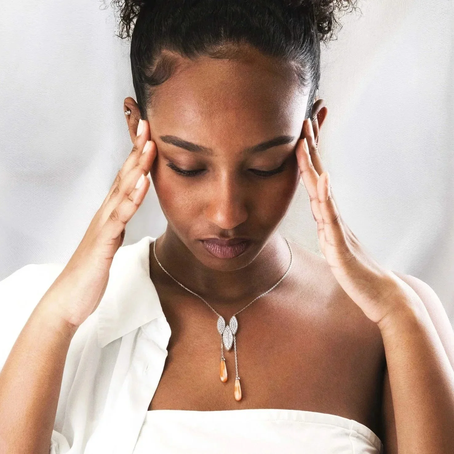 Une femme noire avec des cheveux bouclés, portant des bijoux en or et en argent, avec ses yeux fermés et touchant ses tempes, habillée en blanc.