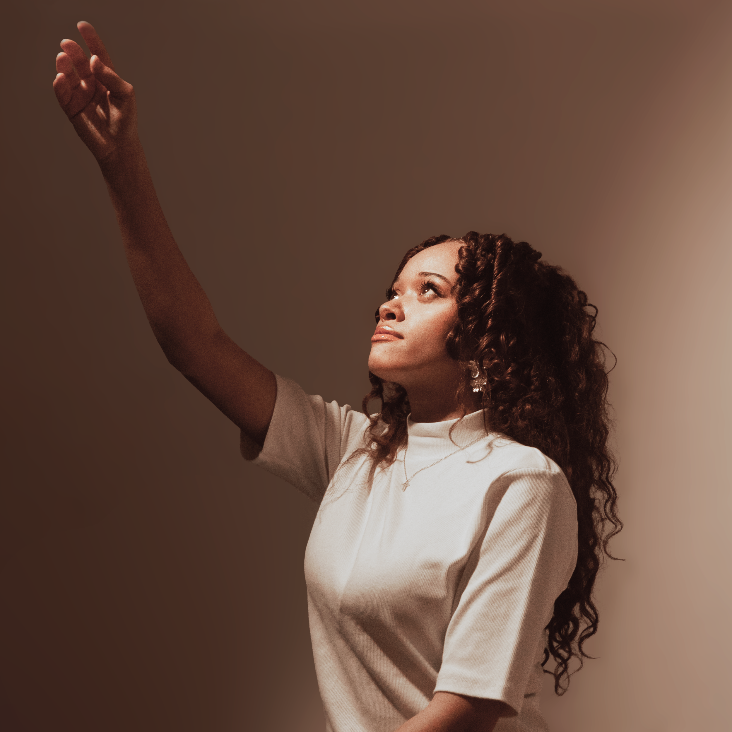 Femme aux cheveux bouclés, regardant vers le haut, portant un t-shirt blanc, bras levé dans un geste artistico-danse.