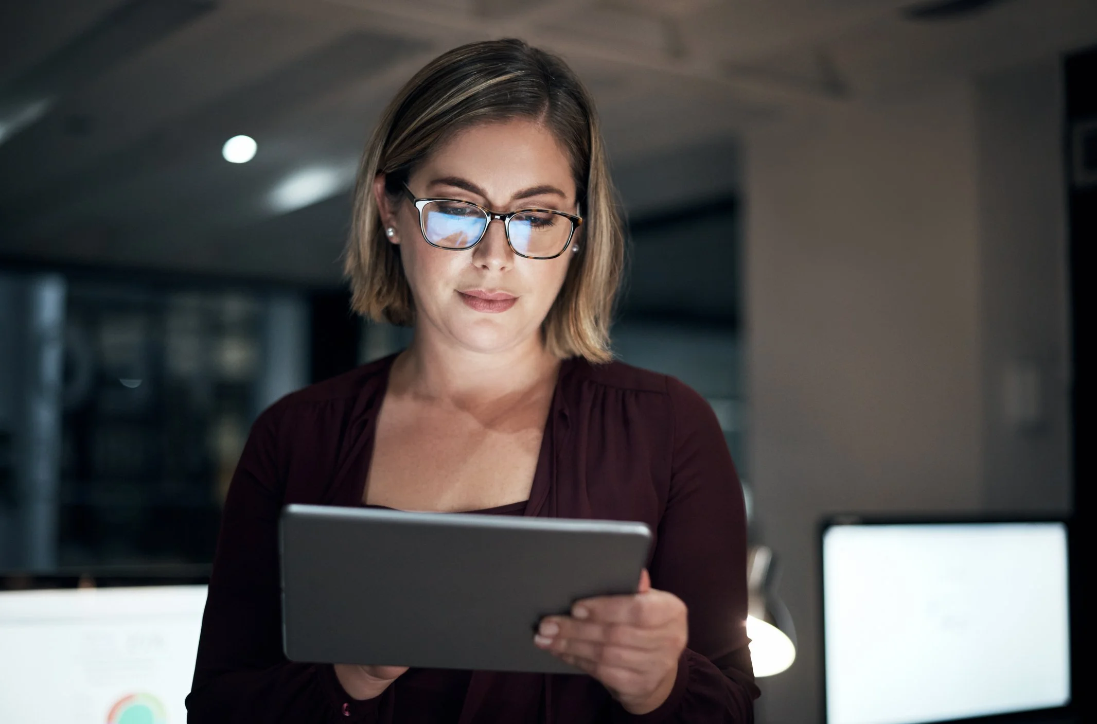A woman with short brown hair, wearing glasses and a maroon top, looks at a tablet computer in a modern indoor setting.