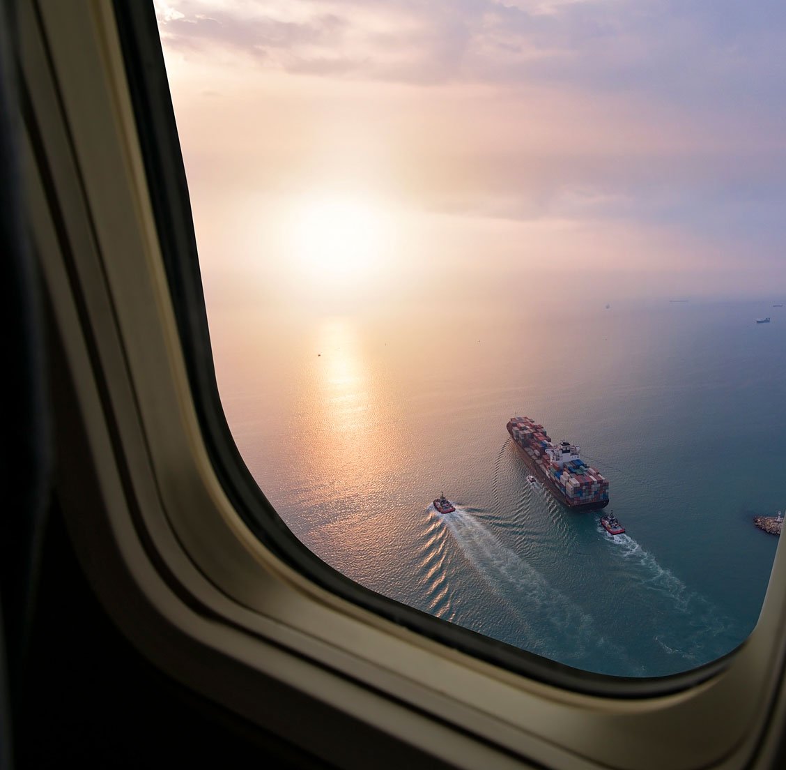 View of the ocean and sky through an airplane window with a cargo ship and two smaller boats visible on the water at sunset.