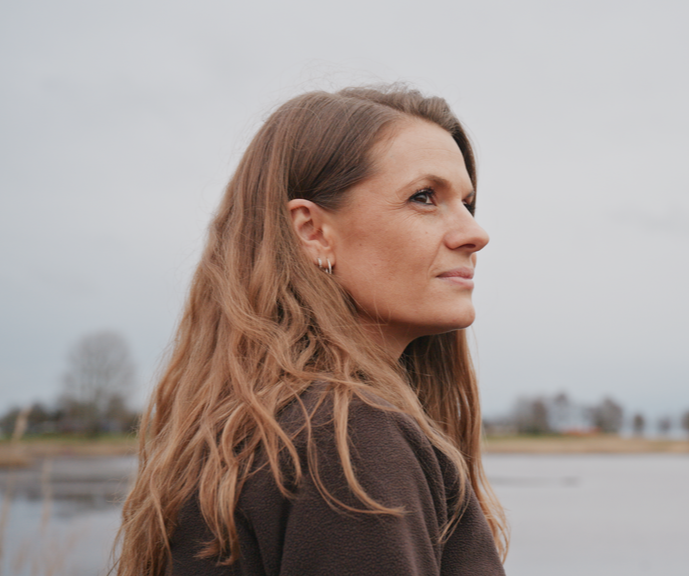 A woman with long, wavy brown hair looking to the right outdoors on a cloudy day, with water and distant trees in the background.
