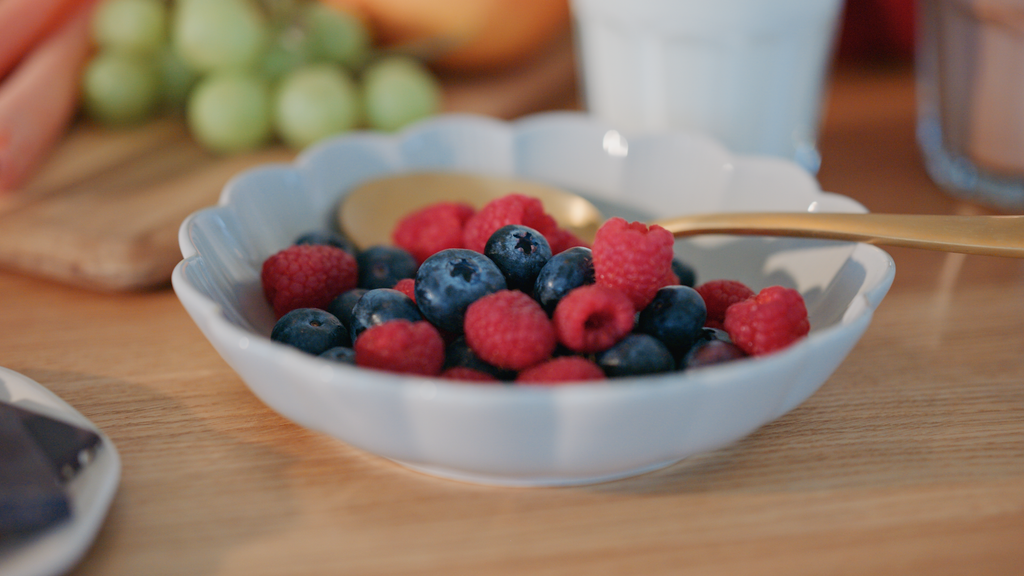 A white bowl filled with blueberries and raspberries on a wooden table, with a gold spoon resting on the bowl's edge.