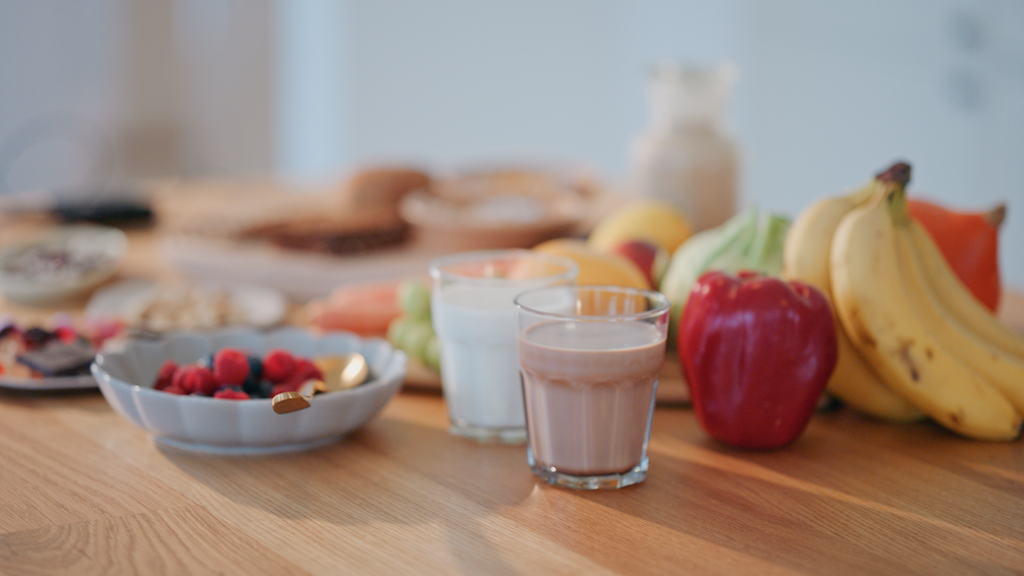 A breakfast spread on a wooden table including a bowl of mixed berries, glasses of milk and chocolate milk, bananas, red apple, grapes, and other breakfast items in the background.