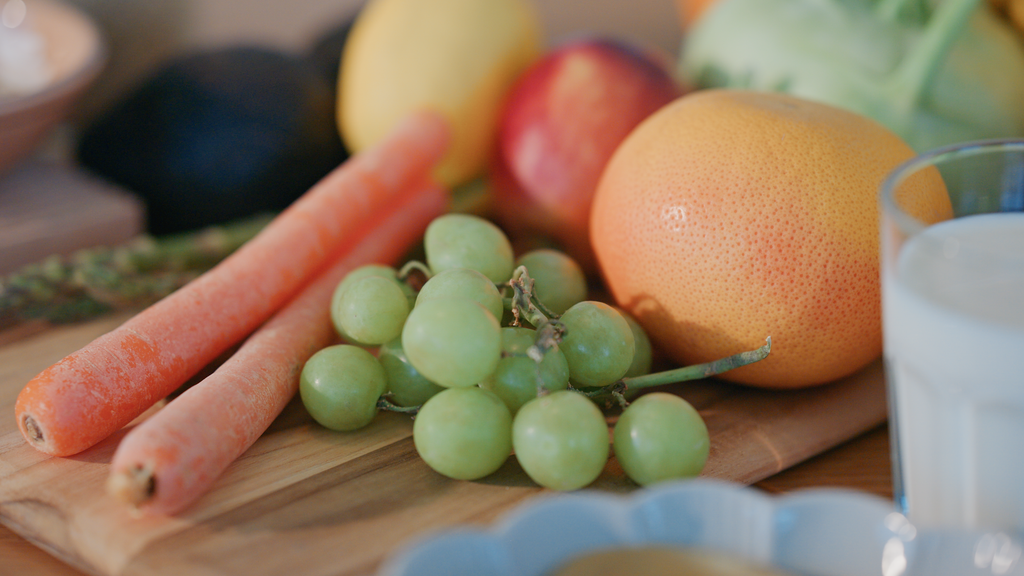 Fresh fruits and vegetables on a wooden cutting board, including carrots, green grapes, an apple, an orange, and a lemon, with a glass of milk nearby.
