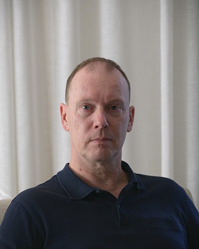 A man with short hair, wearing a navy polo shirt, standing in front of a beige curtain.