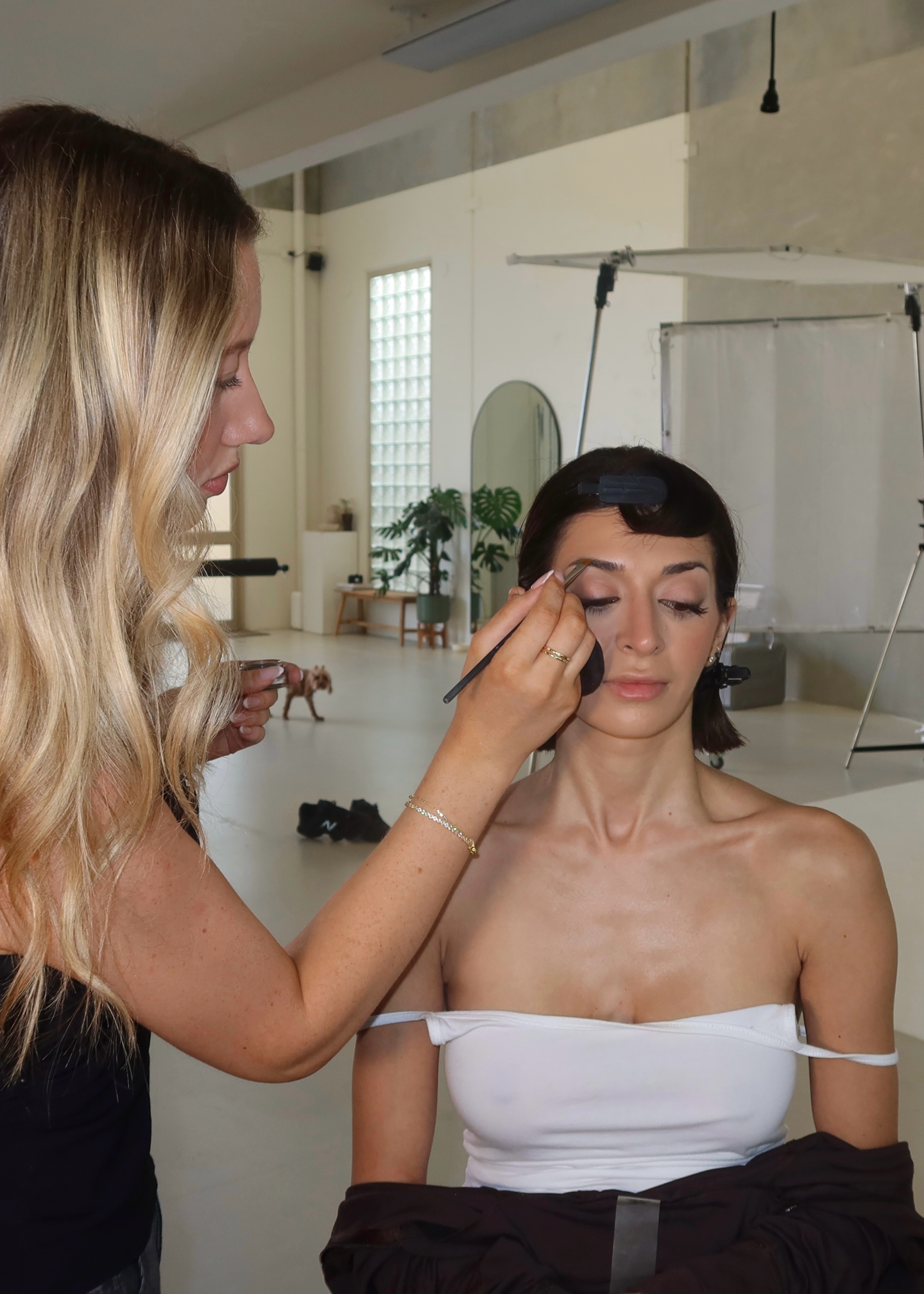 Makeup artist applying eyeshadow to a woman with short dark hair, in a bright studio.