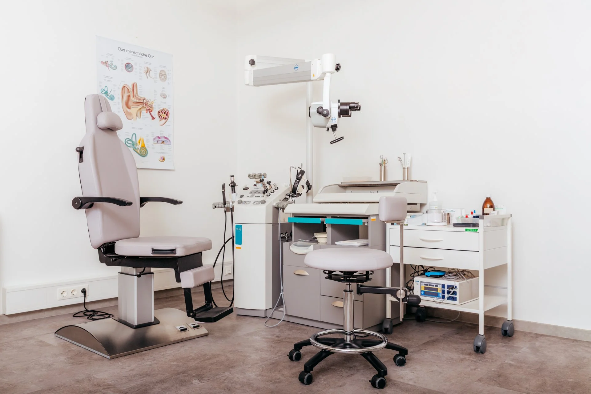 Ein medizinischer Untersuchungsraum mit einem Spezialstuhl auf der linken Seite, einem Labortisch mit medizinischen Instrumenten und Geräten, sowie einem weißen Schrank mit Medikamenten und medizinischen Utensilien auf der rechten Seite.