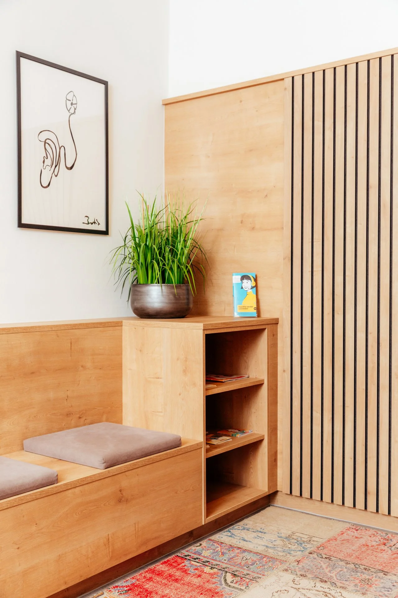 Ein Sitzbereich mit einem hölzernen Sitzbank mit grauen Kissen, einem grünen Pflanzentopf und einer kleinen Bücherregal. An der Wand hängt ein modernes Kunstwerk mit einer schwarzen Linienzeichnung.