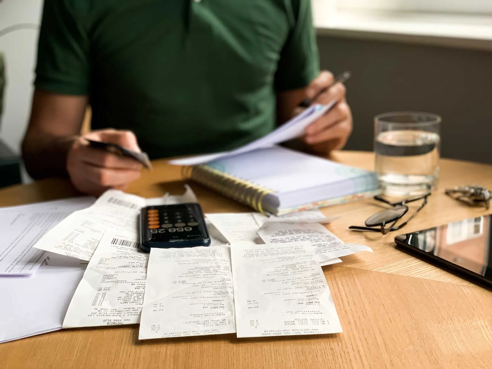 Person in green shirt sitting at a wooden table with receipts, a calculator, glasses, a phone, a notepad, a glass of water, and a tablet, working on financial documents.