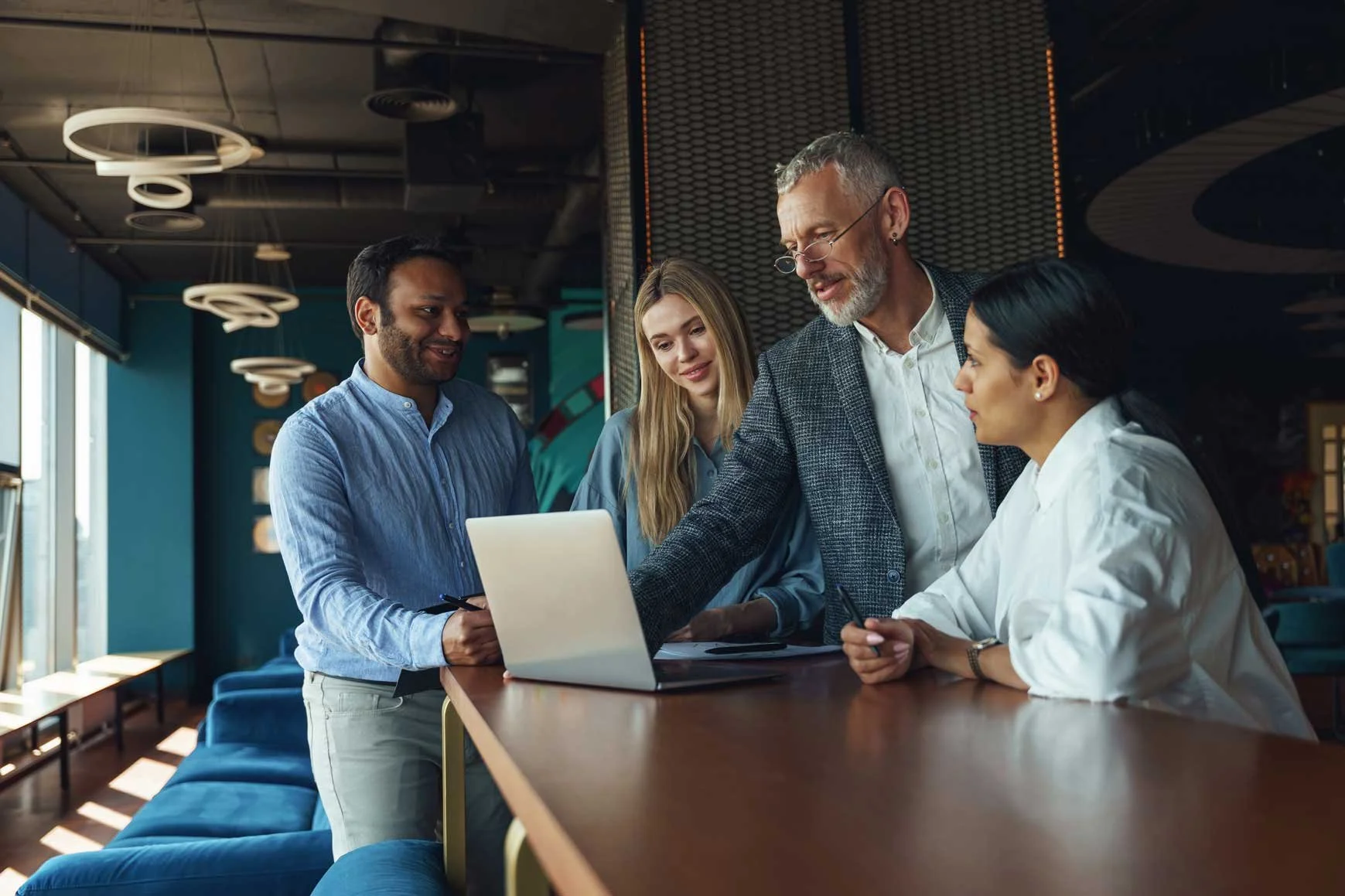Four diverse professionals gathered around a laptop at a modern office counter, engaged in a discussion and analyzing documents together.