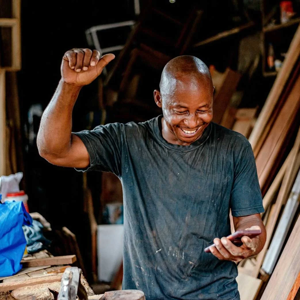 A man with a shaved head smiling and raising one arm while looking at his phone in a woodworking shop or carpentry workspace.