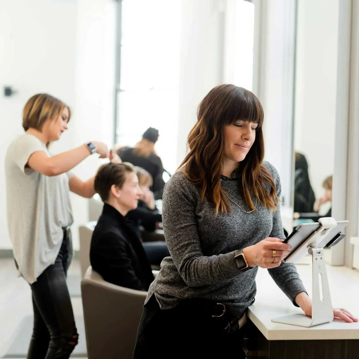 Women working as hairstylists in a salon, with one stylist cutting a client's hair and another standing, using a tablet.