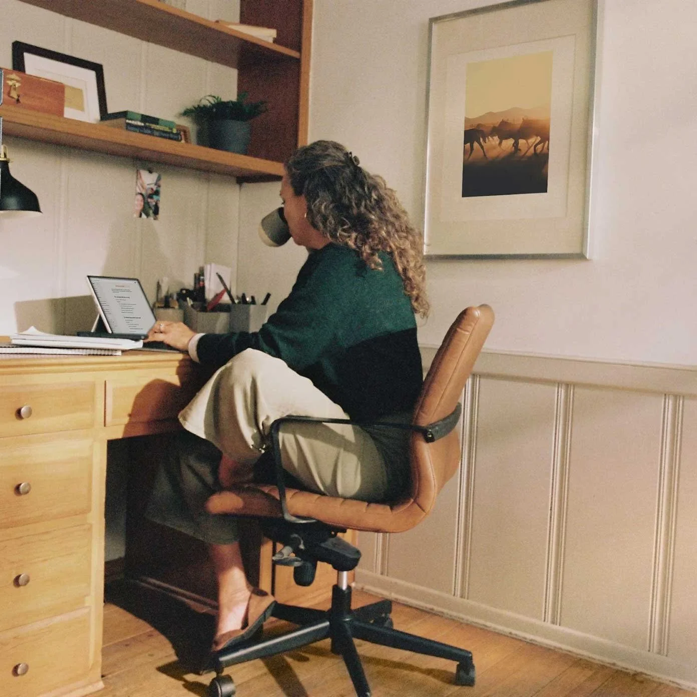 A woman sitting at a wooden desk working on a tablet while drinking from a mug in a home office.