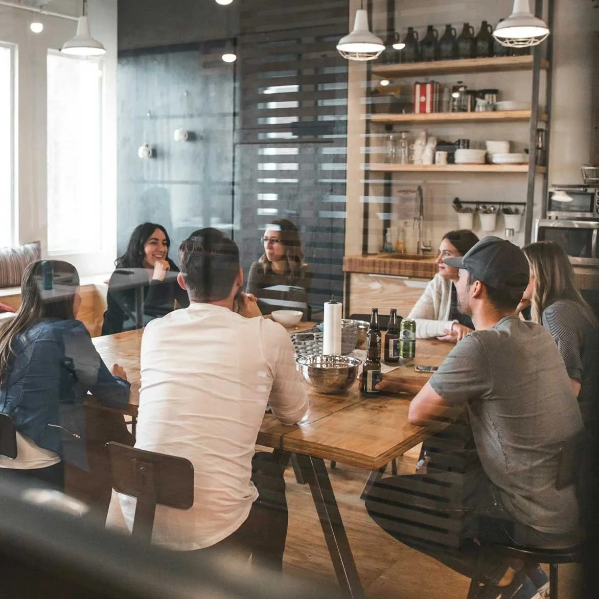 A group of seven people sitting around a wooden table in a cozy, modern cafe, engaged in conversation, with drinks and bowls on the table, viewed through a glass partition.