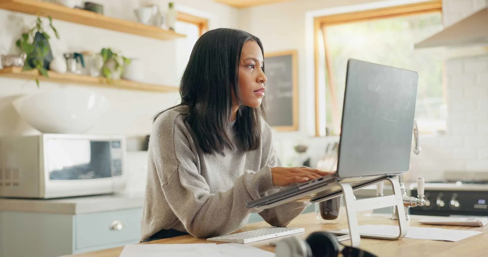 A woman sitting at a wooden kitchen counter working on a laptop with a stand, with a wireless keyboard and papers nearby. The kitchen has open shelves with plants and kitchenware, a microwave, and large windows in the background.