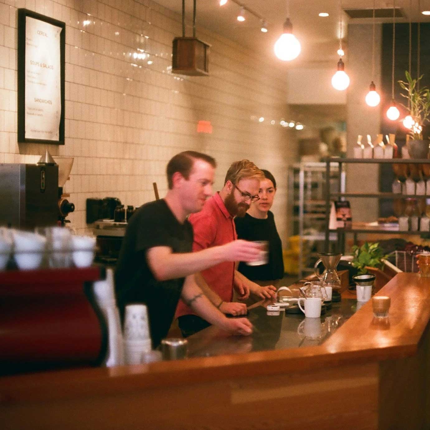 Three people standing behind a wooden bar counter inside a cozy cafe or coffee shop, with hanging pendant lights overhead, a white tiled wall in the background, and shelves with bottles and plants.