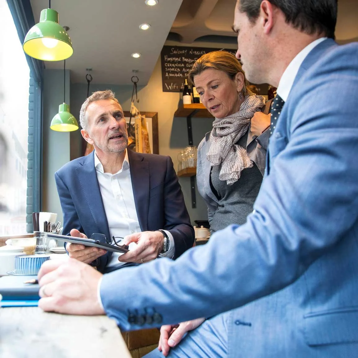 Three people engaged in a conversation at a cafe table, two men and a woman, with menus and drinks in front of them.