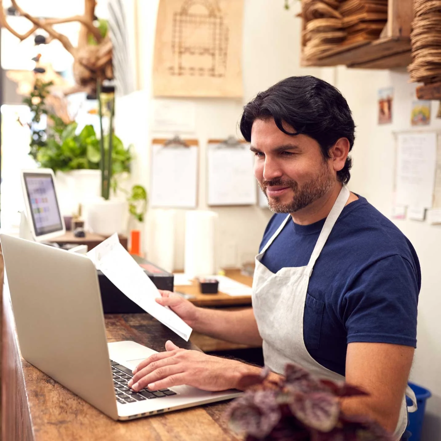 A man wearing a navy blue shirt and a beige apron working at a coffee shop counter, looking at a paper in one hand and using a laptop with the other, with plants and menu boards in the background.