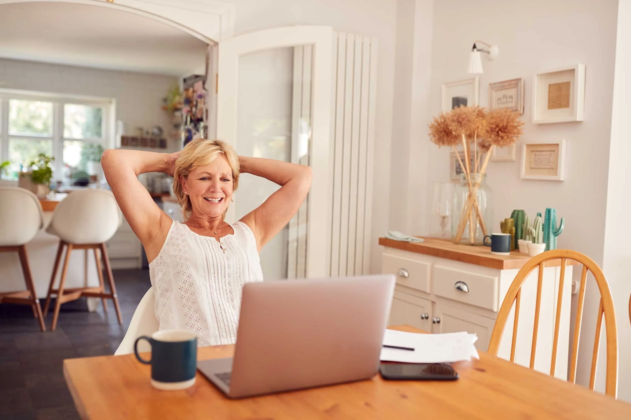 A woman with short blonde hair sitting at a wooden table with a laptop, coffee mug, and papers, smiling and stretching her arms behind her head in a cozy, well-lit kitchen.