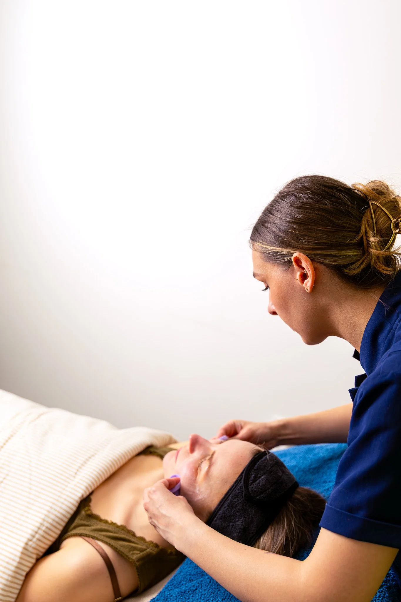Person receiving a facial treatment lying down with a towel over their chest, while a practitioner in a blue uniform and hair clip performs the treatment in a clinical or spa setting.