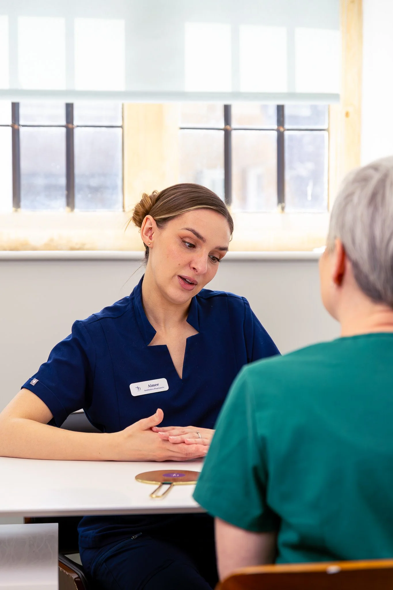 A young woman in navy scrubs with a name tag that reads "Aimee" is having a conversation with an older person wearing a green shirt. They are seated at a white table in a well-lit room with large windows.