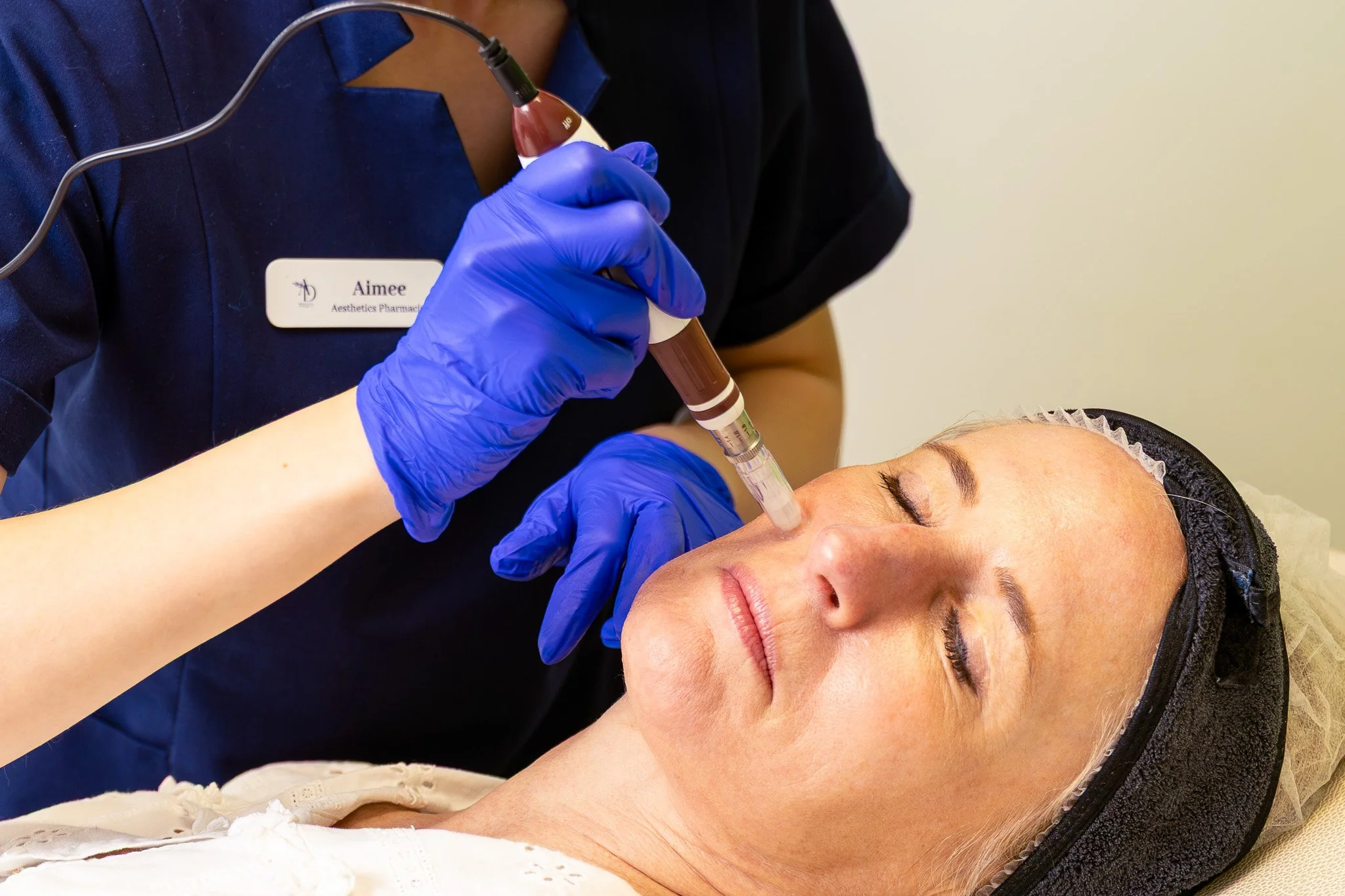 Aesthetician administering a facial treatment with a syringe to a woman lying down with eyes closed, wearing a hair net, in a clinical setting.