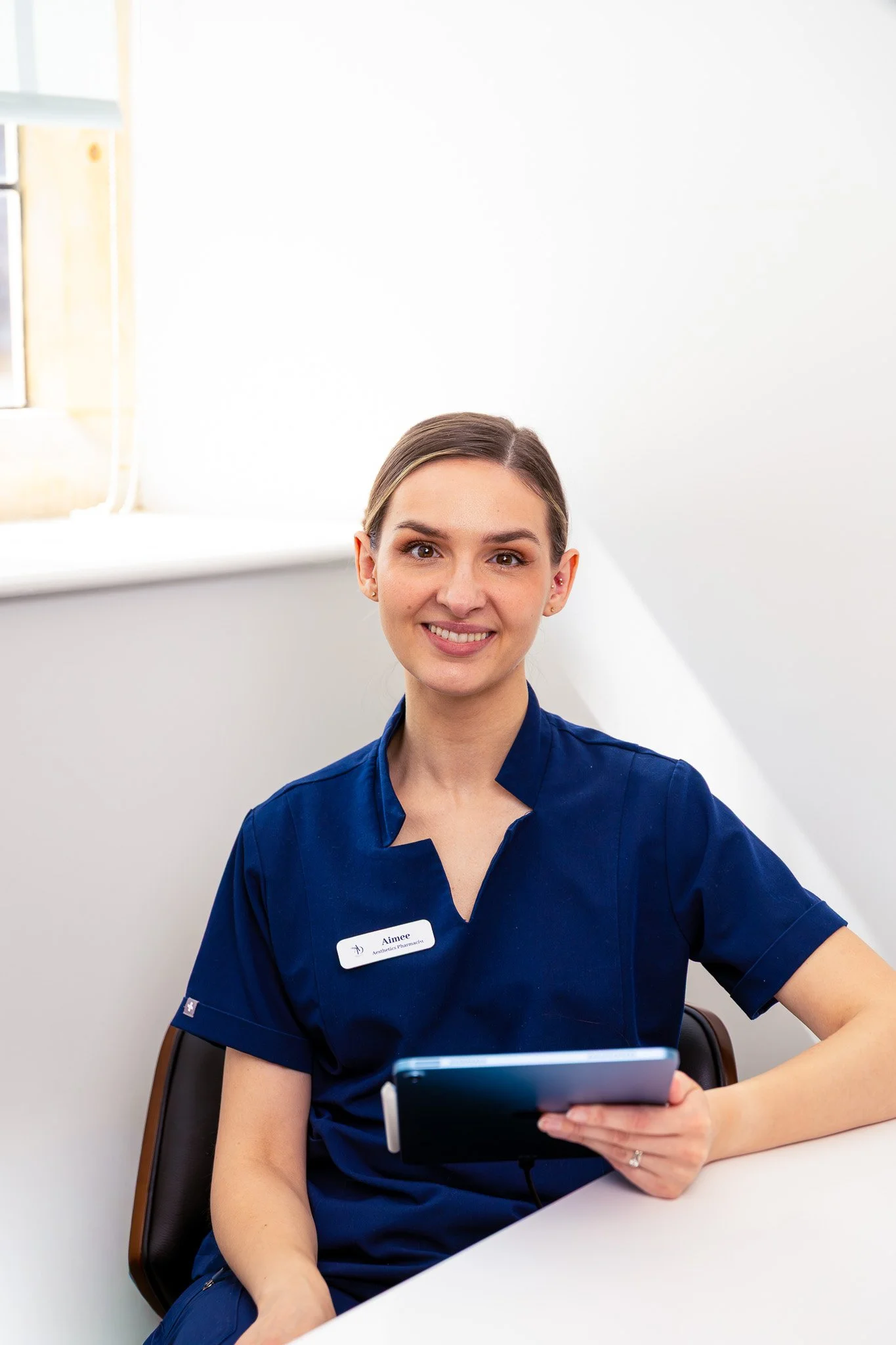 A young woman with brown hair in a bun wearing blue medical scrubs looks at a tablet, sitting in a chair in a bright, white room.
