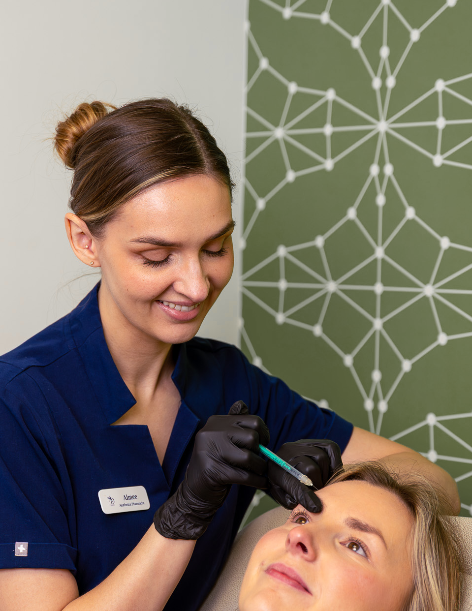 A woman receiving a cosmetic injection in her forehead from a healthcare professional with a name tag that reads 'Aimee,' in a clinic with geometric patterned wall art in the background.