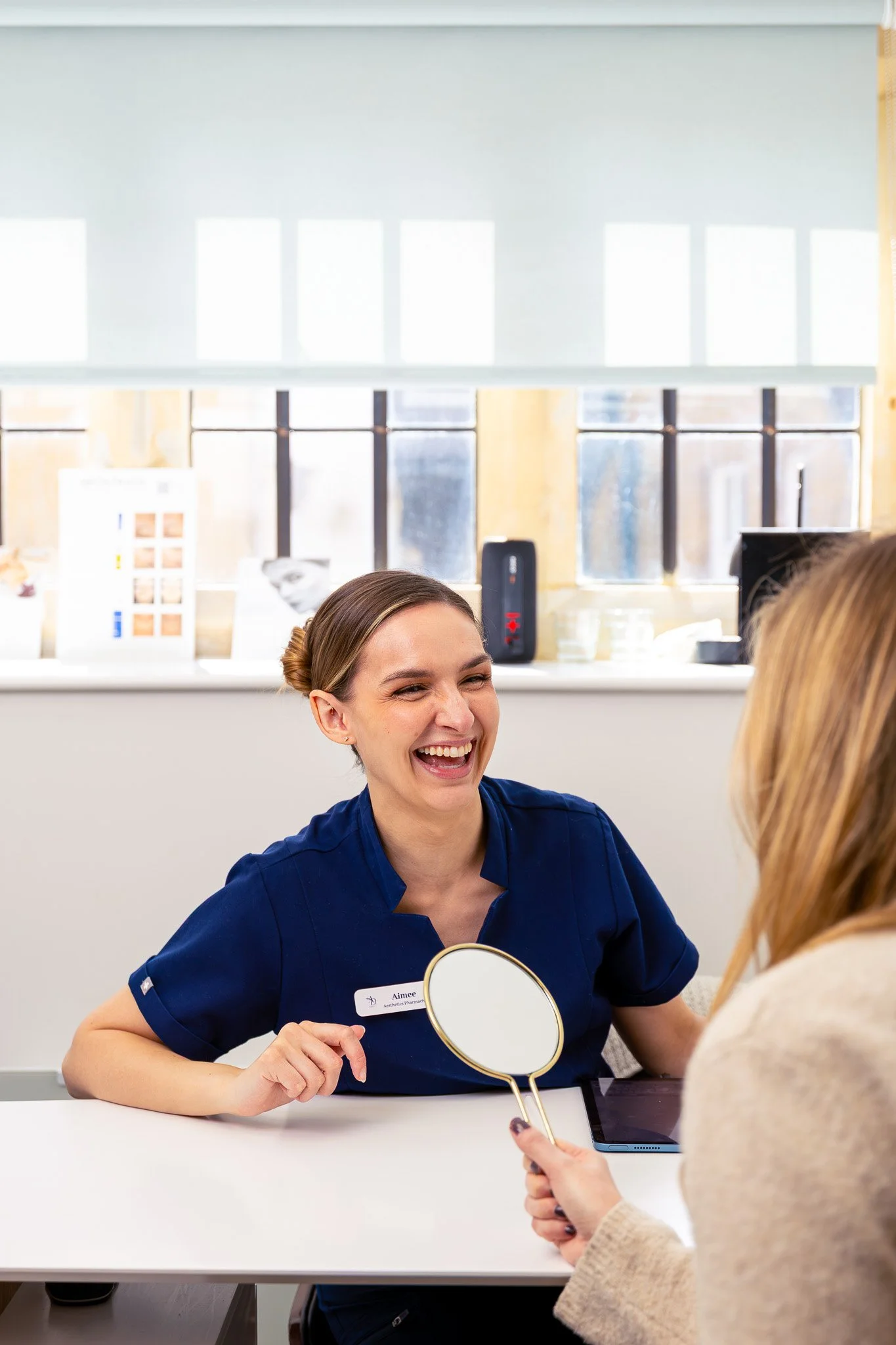 A woman wearing a navy medical uniform sitting at a white table, smiling and holding a round mirror, engaged in a conversation with another woman in a beige sweater, in a well-lit office or medical setting with windows and posters in the background.