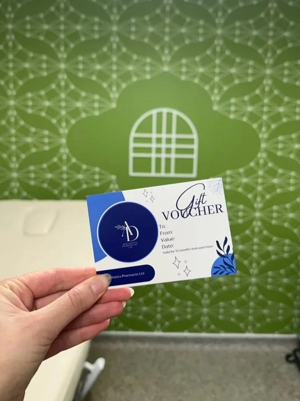 Hand holding a gift voucher with a blue and white design, in front of a green wall with a white stylized window and pattern.