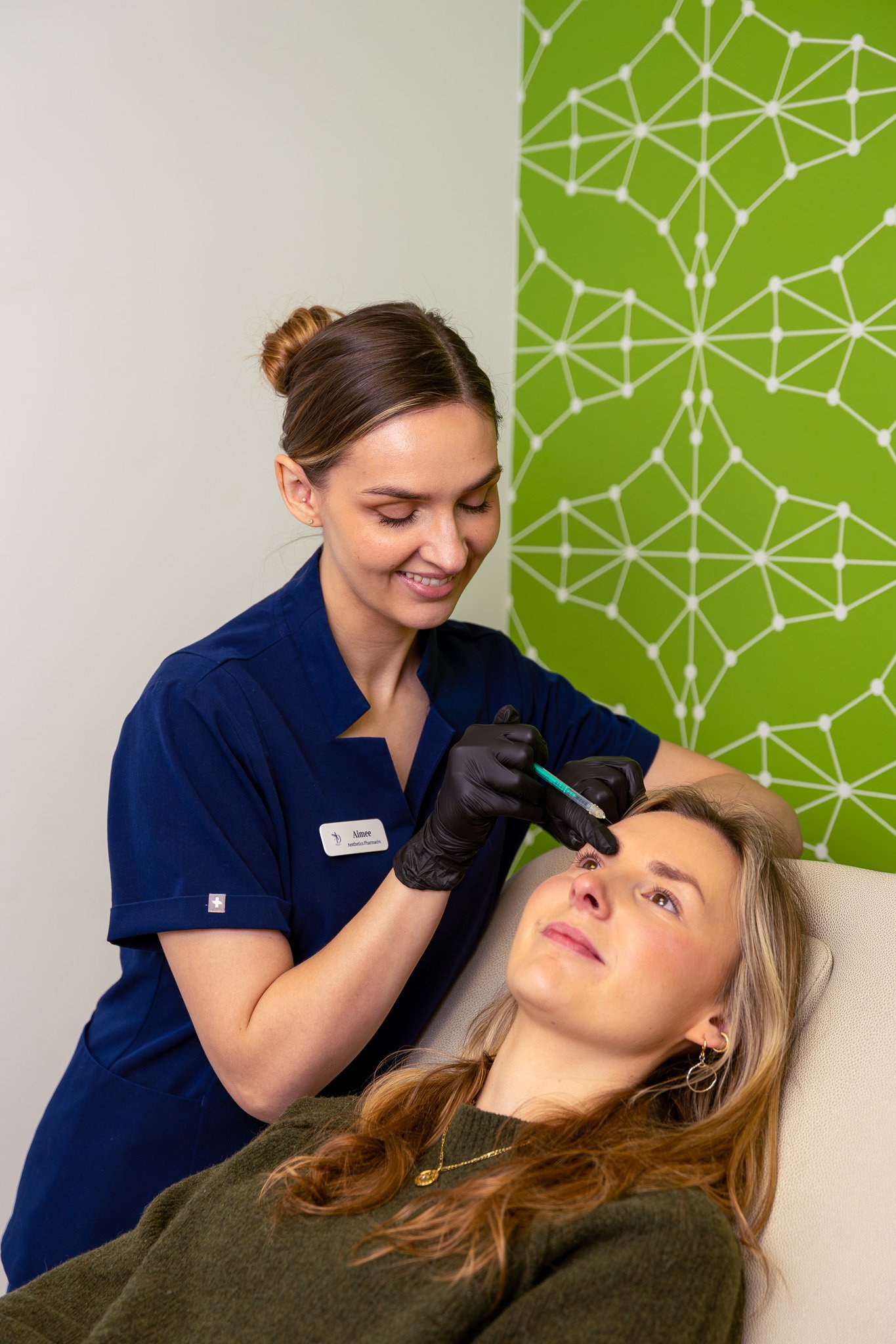 A medical professional administering a cosmetic injection to a woman lying on a medical chair, with a green geometric patterned wall in the background.
