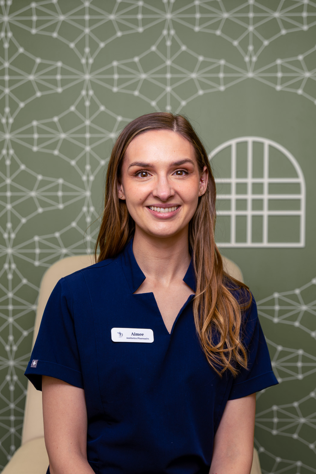 A woman with long brown hair and a blue uniform seated in front of a green wall with a geometric and window design.