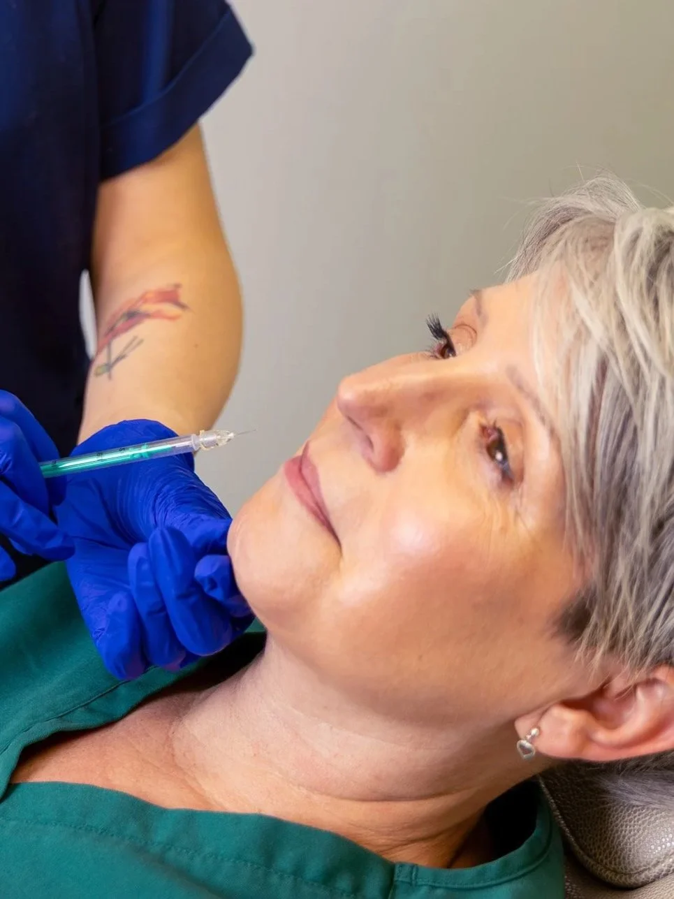 A woman with short gray hair receiving a cosmetic injection in her chin from a healthcare professional wearing blue gloves.