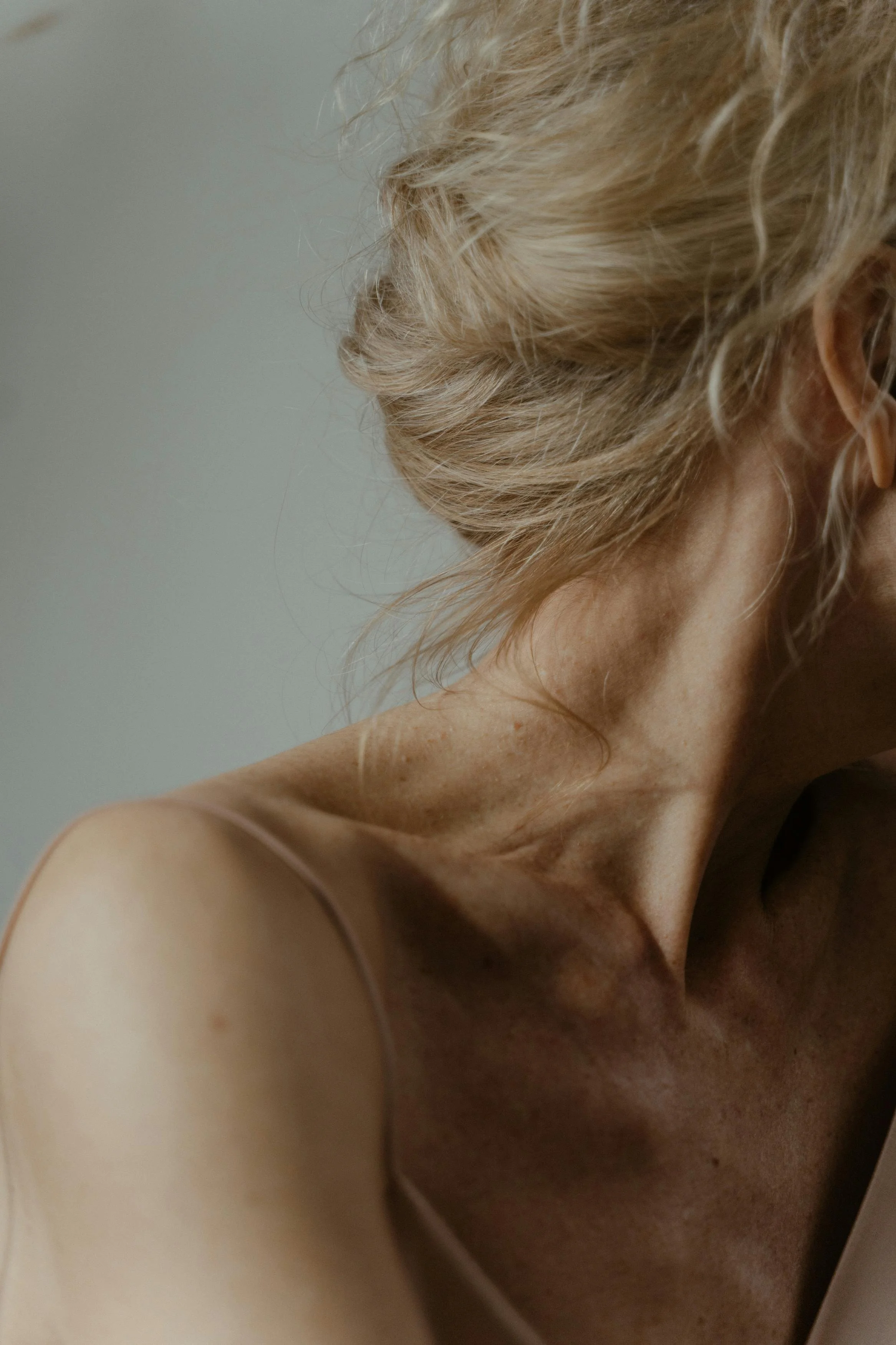 Close-up of a person with blonde curly hair and light skin, wearing a light spaghetti strap top, showing part of their shoulder, neck, and ear.