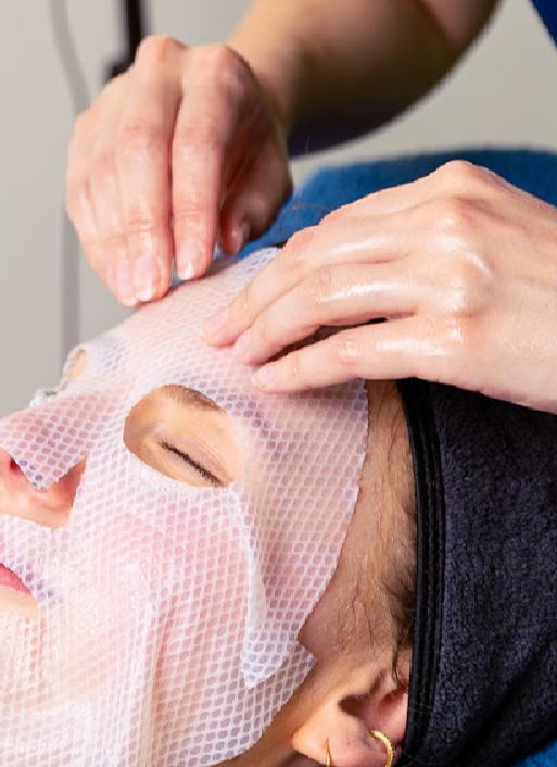 Person receiving a facial treatment with a sheet mask, lying down with eyes closed, in a skincare clinic.