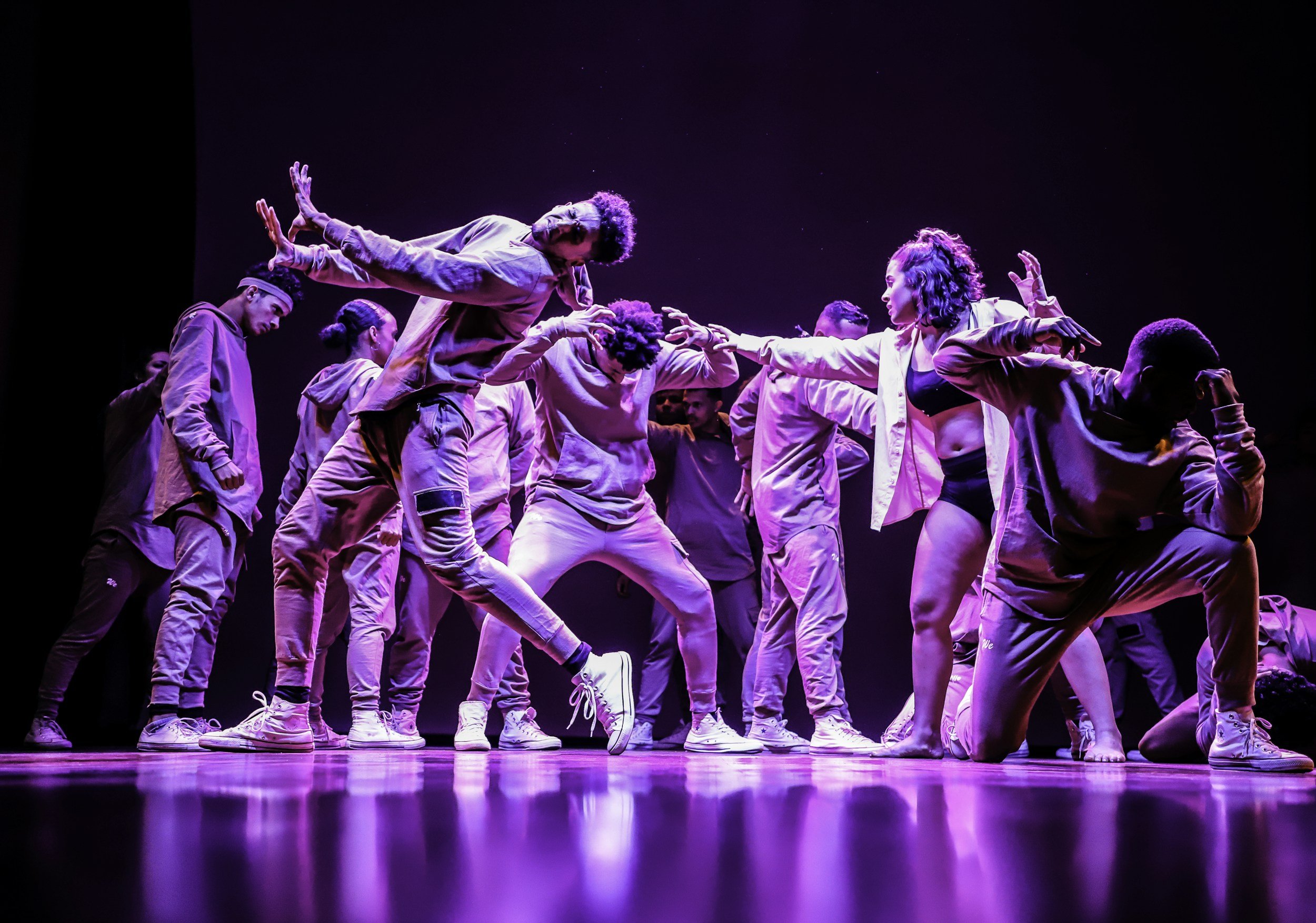 Group of dancers performing on stage with purple lighting, some in athletic clothing, engaging in contemporary dance moves.