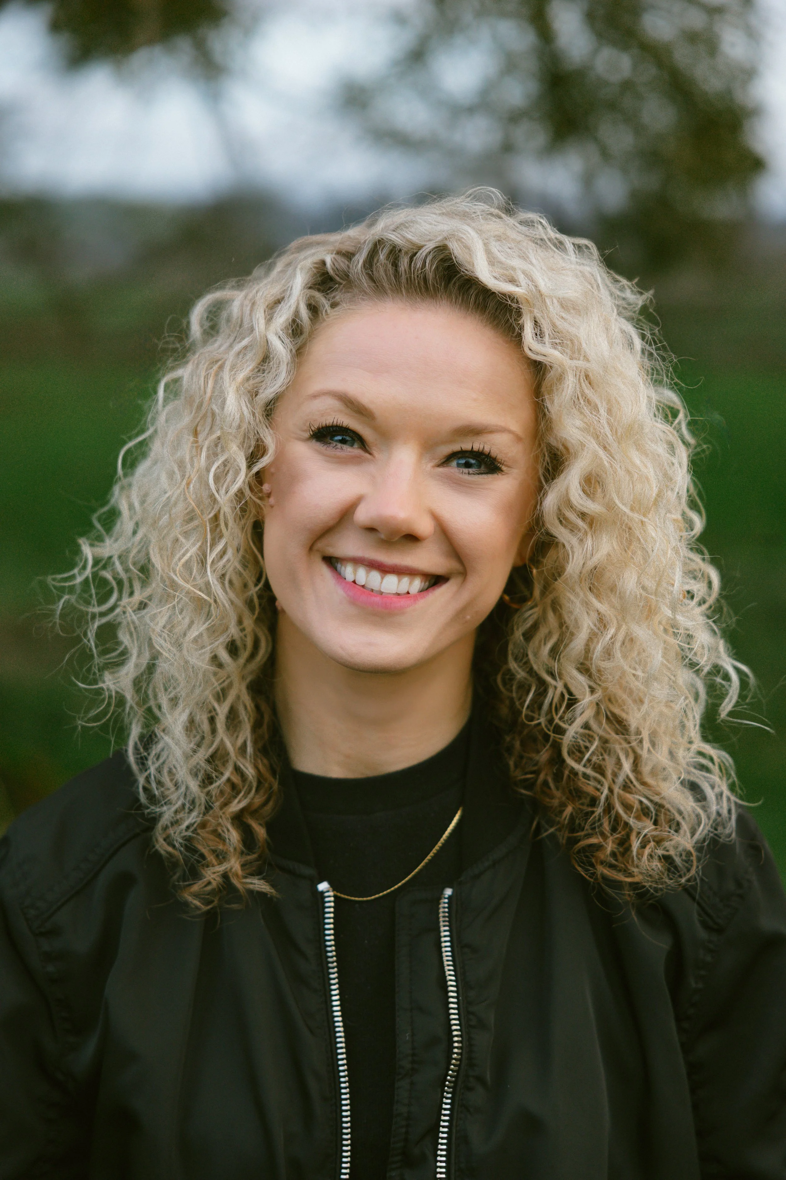 A smiling woman with curly blonde hair outdoors, wearing a black jacket and a gold necklace.