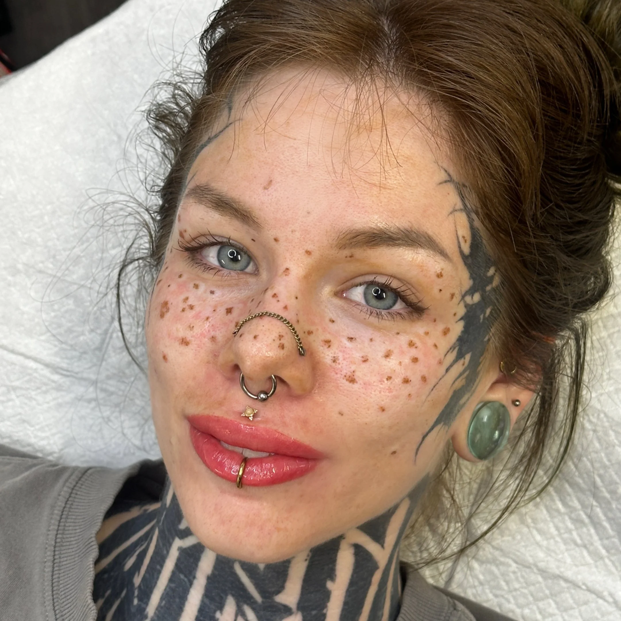 Close-up of a young woman with blue eyes, multiple facial piercings, extensive tattoos on her face and neck, and freckles. Fresh Lip Tattoo, Fresh Freckle tattoo, healed eyebrow tattoo 