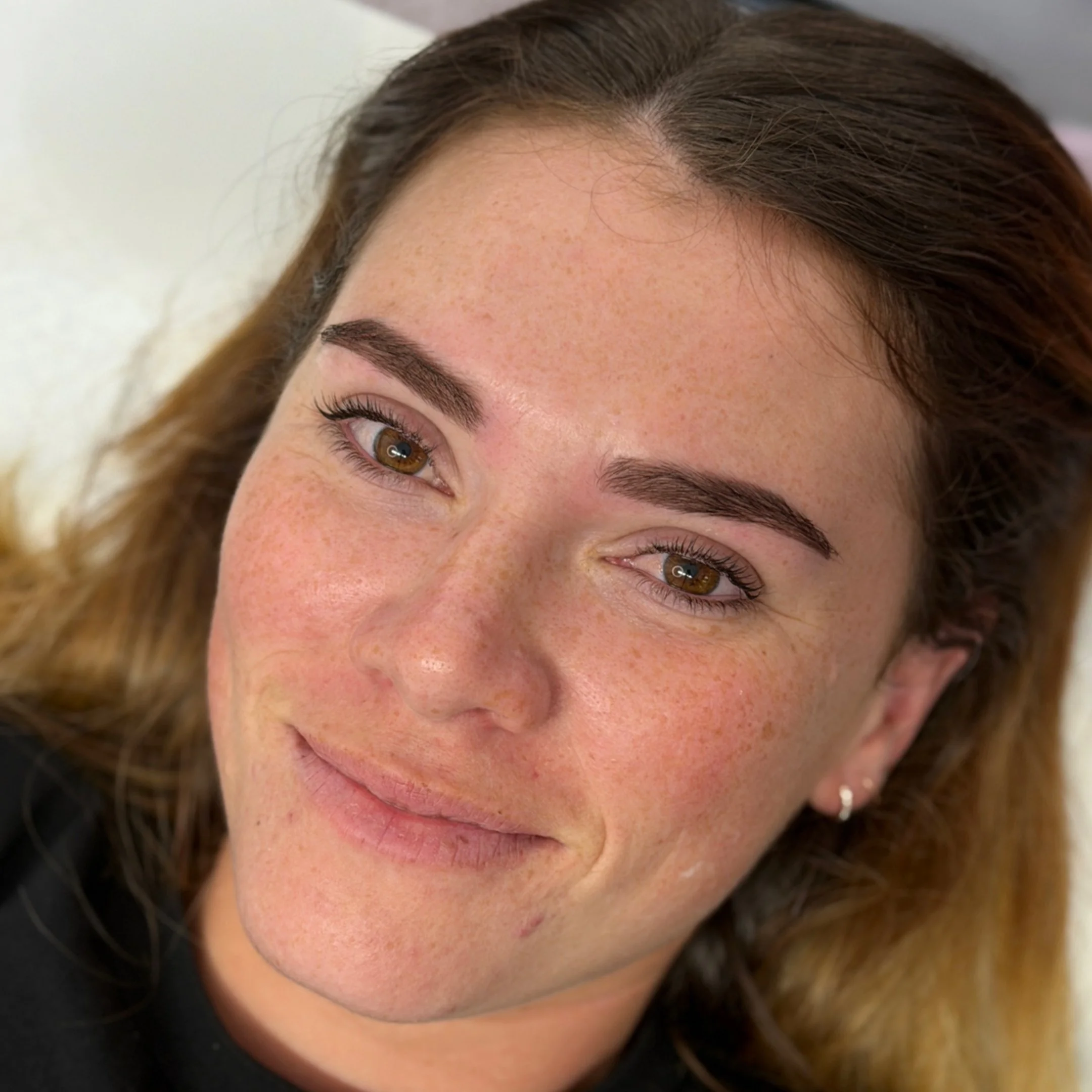 Close-up of a woman with brown hair, brown eyes, and freckles, smiling gently, wearing a small hoop earring. Fresh Brow tinting